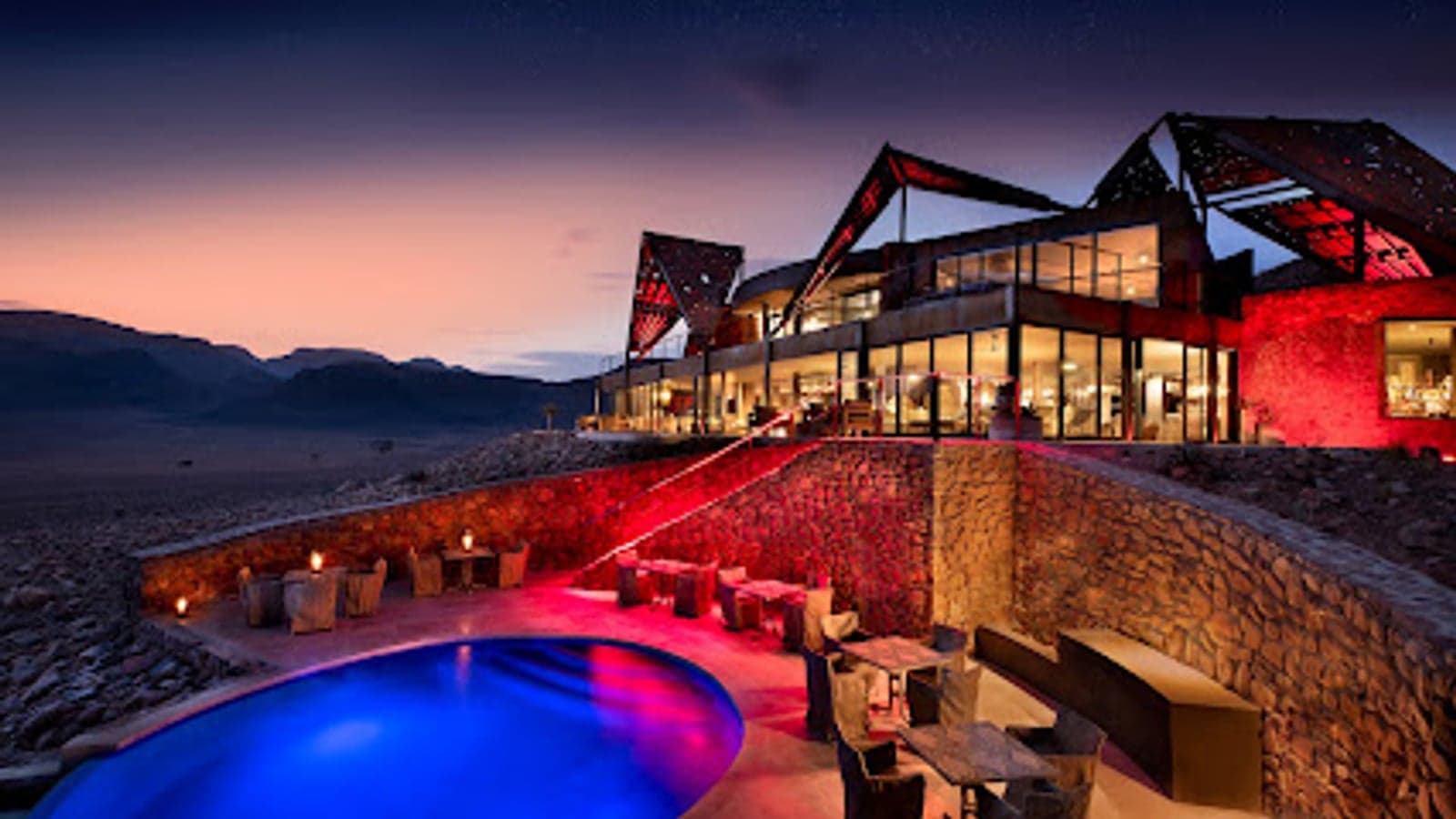 Glass-fronted rooms of Sossusvlei Desert Lodge lit red at night under a starry sky